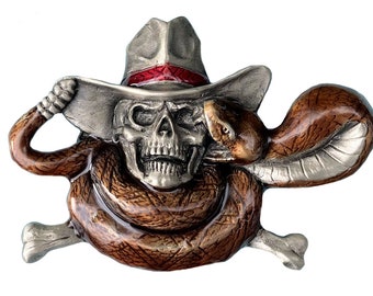 Peut inclure: Boucle de ceinture en métal argenté représentant un crâne portant un chapeau de cowboy avec une bande rouge. Un serpent brun s'enroule autour du crâne et du chapeau, avec des tibias croisés en bas. La boucle a un design détaillé et texturé.