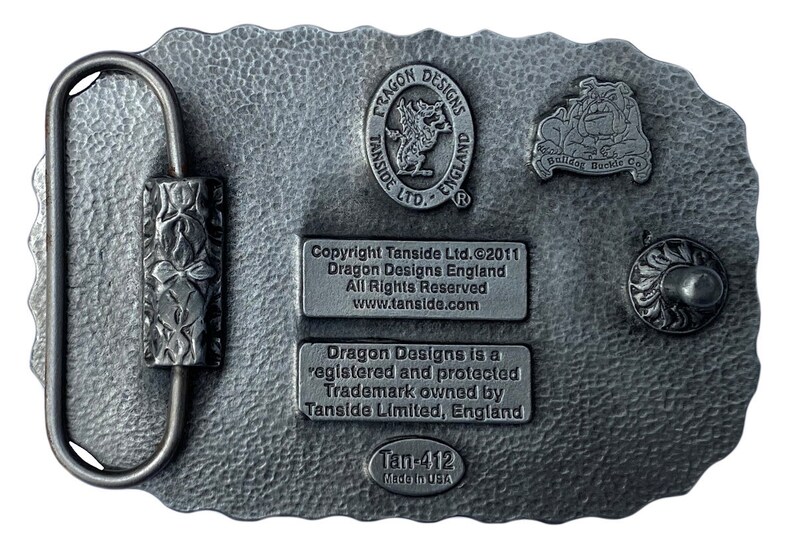 May include: A rectangular, silver-colored belt buckle with a textured surface. The buckle features a loop and a decorative clasp. The back of the buckle has several embossed logos and text, including "Dragon Designs England" and "Bulldog Buckle Co."