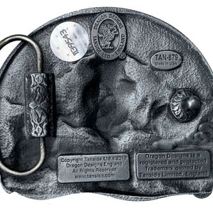 May include: A silver belt buckle with a dragon design. The buckle has a textured surface and a black background. The buckle is labeled "Dragon Designs" and "Tanside Ltd." The buckle also has the text "TAN-879 Made in USA".