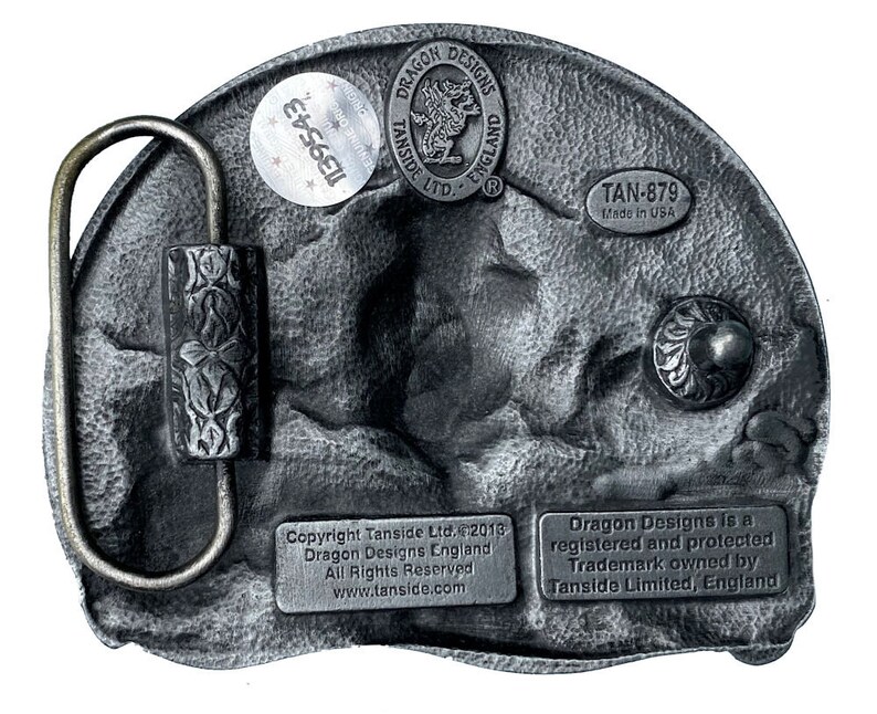 May include: A silver belt buckle with a dragon design. The buckle has a textured surface and a black background. The buckle is labeled "Dragon Designs" and "Tanside Ltd." The buckle also has the text "TAN-879 Made in USA".