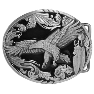 May include: An oval-shaped silver-toned belt buckle featuring a raised relief of a flying eagle in the center. The background is black with intricate silver-toned scrollwork and leaf patterns surrounding the eagle, with two feathers hanging on the right side.