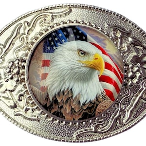 May include: Oval silver-colored belt buckle featuring a detailed image of a bald eagle with the American flag in the background. The eagle has white feathers, a yellow beak, and brown feathers. The buckle has an ornate floral design.