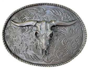 Puede incluir: Hebilla de cinturón de plata con un diseño ornamentado que presenta un cráneo de toro en el centro. El cráneo está detallado con cuernos, ojos y nariz.