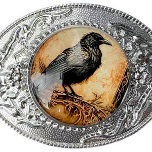 May include: An oval silver-tone belt buckle featuring an embossed floral and vine border. The center contains a circular glass cabochon displaying a detailed illustration of a black raven perched on a branch against a warm, tan-colored background.
