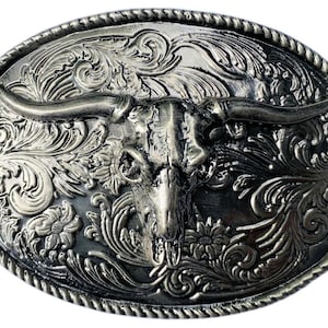 May include: An oval-shaped silver-toned belt buckle featuring a raised steer skull in the center. The background is decorated with intricate, embossed floral scrollwork patterns. The outer edge of the buckle is finished with a twisted rope border.