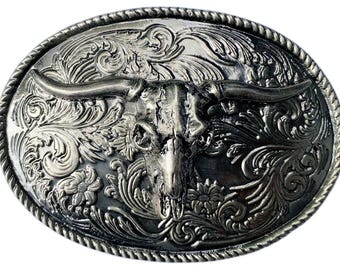 Puede incluir: Una hebilla de cinturón ovalada de tono plateado que presenta un cráneo de toro en relieve en el centro. El fondo está decorado con intrincados patrones de volutas florales en relieve. El borde exterior tiene un acabado de cuerda trenzada.