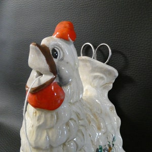 Könnte beinhalten: Weiße Keramik-Hahnfigur mit rotem Kamm und Kehllappen. Der Hahn hat ein braunes Auge und hält eine Garnrolle im Schnabel. Der Hahn ist an einer Wand montiert und hat einen Metallring zum Aufhängen.
