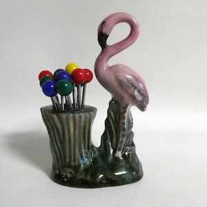 May include: A pink ceramic flamingo figurine with a green base, holding a set of colorful toothpick holders. The toothpick holders are red, blue, green, and yellow.