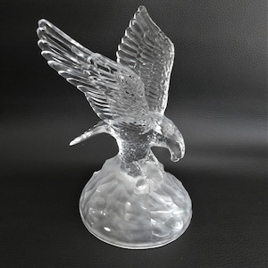 Könnte beinhalten: Eine klare Glasskulptur eines Adlers mit ausgebreiteten Flügeln. Der Adler sitzt auf einem strukturierten Sockel.