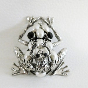 May include: A silver frog brooch with black and white pearl accents. The frog is sitting on its hind legs with its front legs raised. The brooch is a statement piece that would add a touch of whimsy to any outfit.