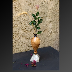 May include: A wooden vase with a dark brown and light wood grain pattern, with a small base and a wide opening. The vase is filled with a sprig of green leaves and a small pink flower. The vase is sitting on a white marble base.