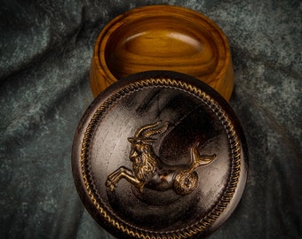 May include: A wooden box with a dark brown lid featuring a gold-colored zodiac symbol of a sea goat. The box is open, revealing a light brown wooden interior.