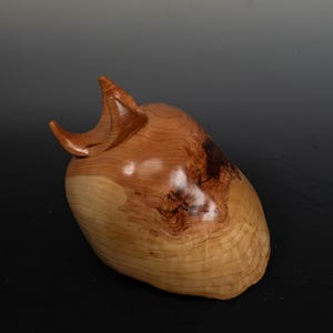 May include: A handcrafted wooden sculpture with a smooth, polished finish. The sculpture is a light brown color with darker brown accents and a unique, organic shape. The piece has a curved, horn-like protrusion on top.