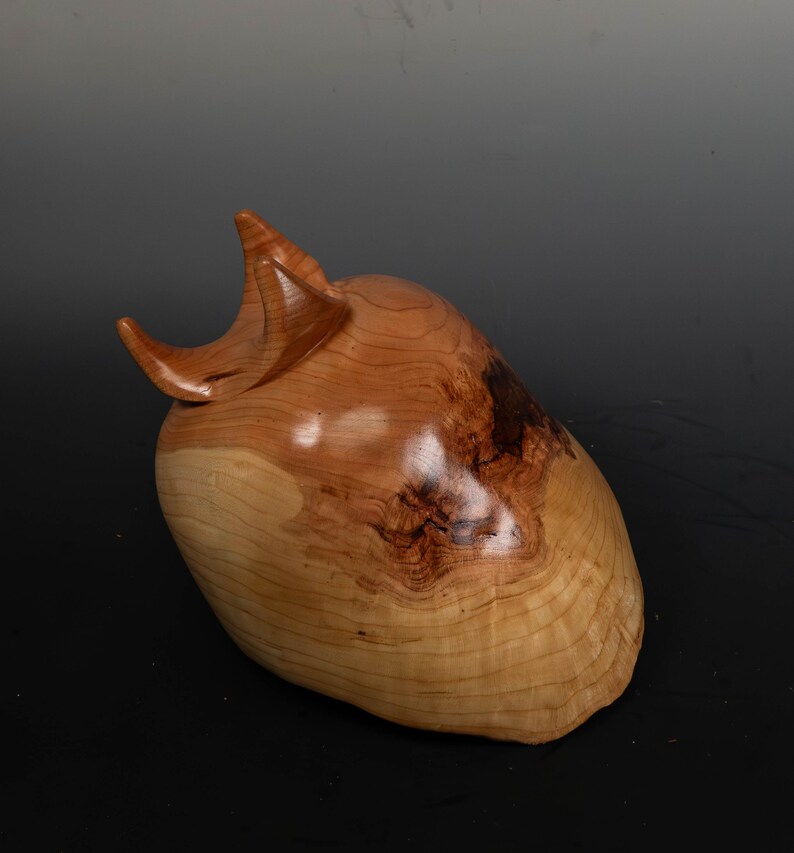 May include: A handcrafted wooden sculpture with a smooth, polished finish. The sculpture is a light brown color with darker brown accents and a unique, organic shape. The piece has a curved, horn-like protrusion on top.