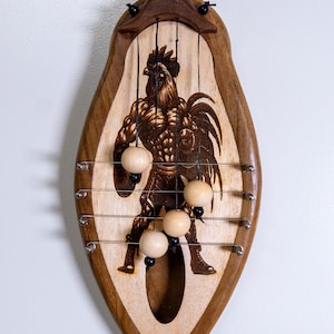 May include: Wooden musical instrument with a rooster design and hanging wooden balls. The instrument is shaped like a teardrop and has a brown wood frame with a light wood centre.