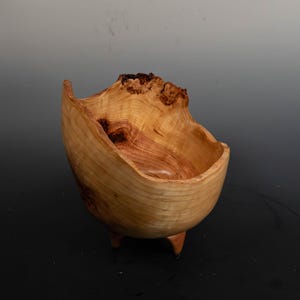 May include: A handcrafted wooden bowl with a natural, organic shape. The bowl is light brown with darker wood grain and sits on three small legs. The rim has a unique, uneven edge, adding to its rustic charm. The bowl is likely made from a single piece of wood.