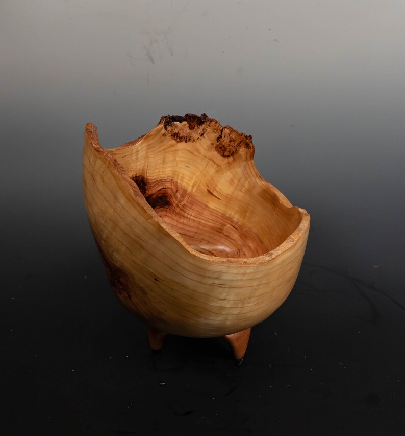 May include: A handcrafted wooden bowl with a natural, organic shape. The bowl is light brown with darker wood grain and sits on three small legs. The rim has a unique, uneven edge, adding to its rustic charm. The bowl is likely made from a single piece of wood.