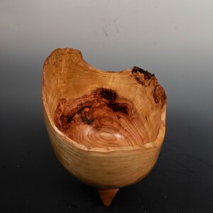 May include: A handcrafted wooden bowl with a natural, organic shape. The bowl features a light brown exterior with darker brown and reddish-brown interior wood grain patterns. It has a unique, asymmetrical rim and a small, pointed base.