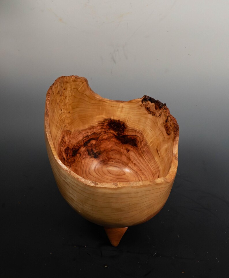 May include: A handcrafted wooden bowl with a natural, organic shape. The bowl features a light brown exterior with darker brown and reddish-brown interior wood grain patterns. It has a unique, asymmetrical rim and a small, pointed base.