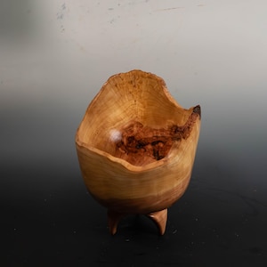 May include: A handcrafted wooden bowl with a natural, organic shape. The bowl is light brown with darker wood grain patterns and rests on three small, curved legs. The rim has an uneven, textured edge.