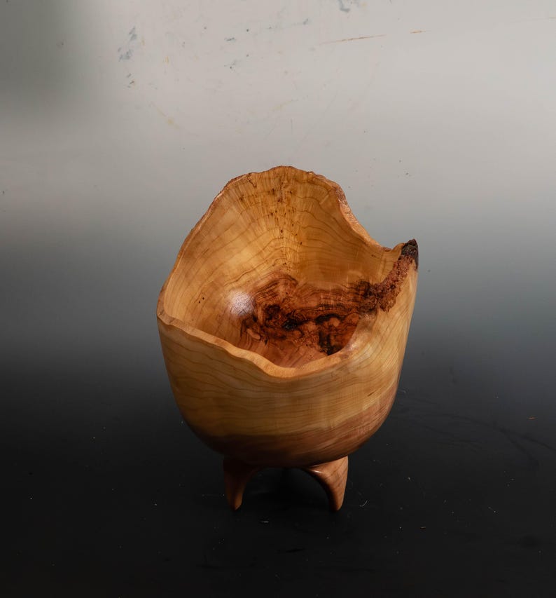 May include: A handcrafted wooden bowl with a natural, organic shape. The bowl is light brown with darker wood grain patterns and rests on three small, curved legs. The rim has an uneven, textured edge.