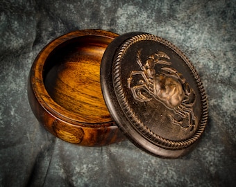 May include: A wooden box with a dark brown lid featuring a carved crab design. The box is open, revealing the interior, which is made of light brown wood.