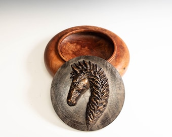 May include: A wooden bowl with a lid featuring a bronze-colored horse head relief. The bowl is made of a light brown wood, and the lid is a circular, dark gray-brown color.