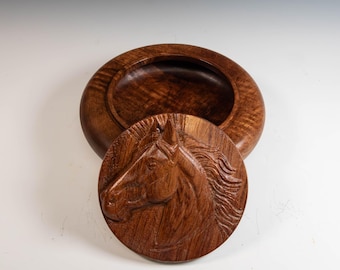 May include: A round, wooden box with a lid featuring a carved horse head. The box and lid are made of a dark brown wood with a visible grain pattern. The lid is circular and fits snugly on top of the box.