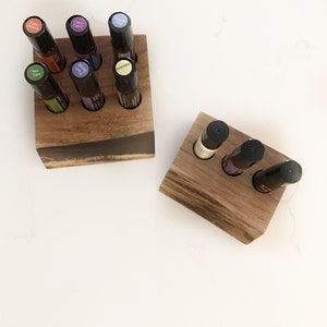 May include: Two wooden essential oil holders, each with holes for small glass bottles. The holders are made of dark wood with a natural grain. The bottles have black caps and colourful labels. The holders are on a white surface.