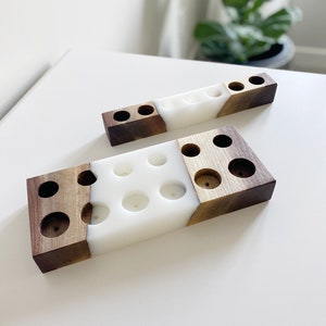 May include: Two wooden candle holders with a white resin centre. The holders are rectangular with dark brown wood on the ends and circular holes for candles. One holder is longer than the other.