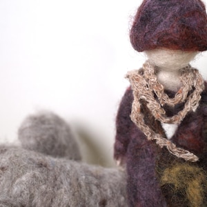 May include: A felt doll with a purple hat and a brown scarf. The doll is wearing a purple and brown patterned dress. The doll is sitting on a gray surface.
