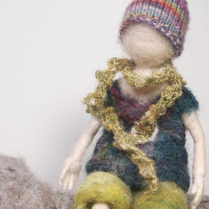 May include: A felt doll wearing a colorful knitted hat and a long, crocheted scarf. The doll has a blue and green patterned dress and is sitting on a gray surface.