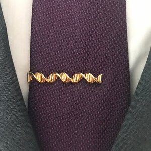 Puede incluir: Un broche de corbata de color dorado en forma de doble hélice, símbolo del ADN, está sujeto a una corbata de color morado oscuro.