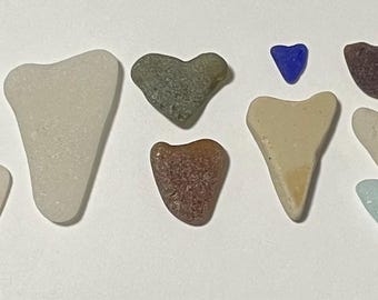 May include: A collection of twenty-one heart-shaped sea glass and ceramic pieces in various colors including white, beige, green, blue, and brown. One white ceramic piece features a small brown floral pattern, and all items are arranged in a loose group.