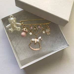 Pu&ograve; includere: Una spilla per neonati in oro con un cavallo a dondolo, un orsacchiotto, un ciuccio, stelle e un fiocco. La spilla &egrave; in una scatola bianca.