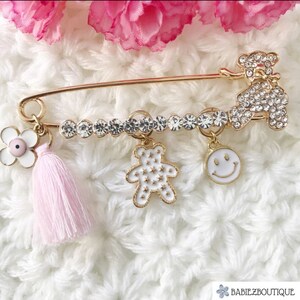 Può includere: Una spilla di sicurezza dorata con un fiocco rosa, un ciondolo a forma di fiore bianco, un ciondolo a forma di orsacchiotto bianco con stelle bianche e un ciondolo a forma di faccina sorridente bianca. La spilla è decorata con strass trasparenti.
