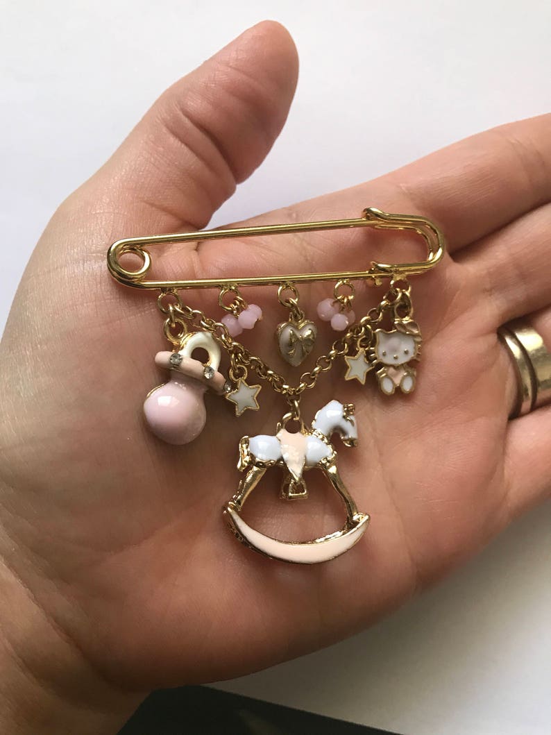 Pu&ograve; includere: Una spilla di sicurezza dorata con ciondoli, tra cui un cavallo a dondolo, un ciuccio, un cuore, una stella e un personaggio Hello Kitty. I ciondoli sono attaccati alla spilla con una catena.