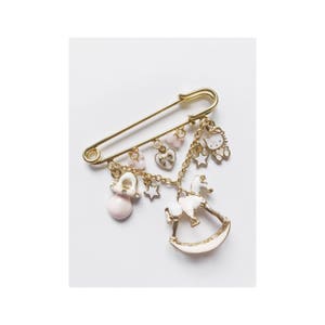 Pu&ograve; includere: Spilla di sicurezza dorata con ciondoli rosa e bianchi, tra cui un cavallo a dondolo, un ciuccio, un cuore, una stella e un personaggio Hello Kitty.