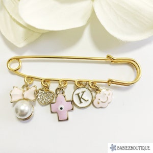 Può includere: Spilla di sicurezza dorata con ciondoli, tra cui un fiocco rosa, un cuore di strass trasparente, una croce rosa con un occhio turco blu, un cerchio bianco con la lettera K e una nuvola rosa con un faccina sorridente.