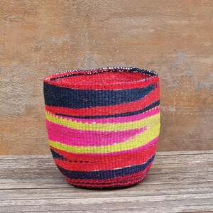 May include: A colourful, handwoven basket with a cylindrical shape. It has horizontal stripes in red, navy blue, pink, and yellow. The rim has a dark trim. The basket is made of natural fibres and sits on a wooden surface.
