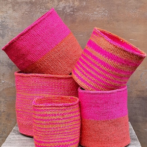 Op de afbeelding: Een verzameling geweven manden in verschillende maten en vormen, voornamelijk roze en oranje. Sommige manden hebben streepjespatronen. De manden staan op een houten oppervlak, waardoor hun texturen en kleuren worden benadrukt.