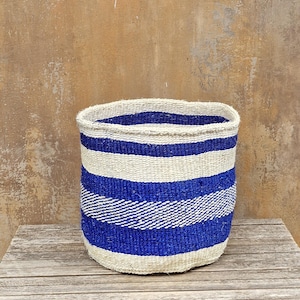 以下が含まれることがあります： 白と青の水平ストライプが交互に配置された織りバスケット。バスケットは円筒形で、上部がわずかに広がっており、木製の表面に置かれています。背景は、テクスチャ加工されたニュートラルカラーの壁です。