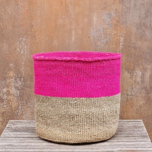 May include: A round woven basket with a bright pink top and a natural beige bottom. The basket is made of tightly woven fibres and sits on a wooden surface. The basket is a decorative storage container.