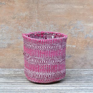 May include: A cylindrical woven basket featuring a repeating pattern of horizontal stripes in vibrant magenta and neutral grey. The textile construction displays a visible herringbone weave texture throughout the exterior surface of the container.