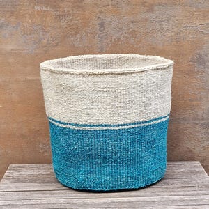 May include: A woven basket with a cylindrical shape. The top half is a natural beige color, while the bottom half is a vibrant teal. A thin white stripe separates the two colors. The basket is made of a textured material and sits on a wooden surface.