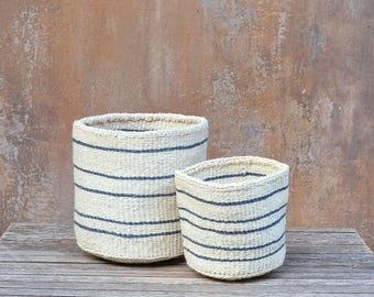 Puede incluir: Dos cestas tejidas cilíndricas hechas de fibra natural de color claro con rayas horizontales de color azul oscuro. Las cestas están abiertas por la parte superior y presentan un patrón de tejido texturizado y consistente en toda su estructura.