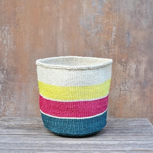 May include: A cylindrical woven basket featuring horizontal colour-blocked stripes. The top section is off-white, followed by a vibrant yellow band, a bright pink middle section, and a deep teal base. The basket has a textured, natural fibre finish.