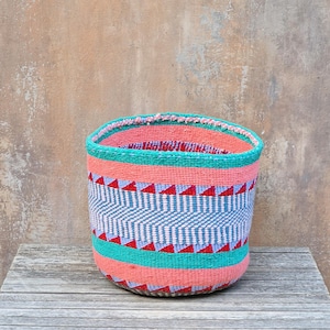 May include: A cylindrical woven basket featuring horizontal bands of coral, turquoise, and a central chevron pattern in light blue and white. The design includes thin rows of red triangles bordering the chevron section, creating a vibrant look.