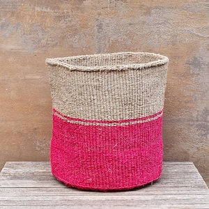 Puede incluir: Una cesta tejida de forma cilíndrica. La mitad superior es de color beige natural, mientras que la mitad inferior es de color rosa vibrante. Una fina franja rosa separa los dos colores. La cesta está sobre una superficie de madera.