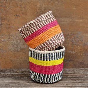 Puede incluir: Dos cestas tejidas cilíndricas apiladas verticalmente. La cesta superior presenta patrones de zigzag en marrón y blanco con bandas horizontales de rosa brillante y naranja. La cesta inferior muestra patrones de zigzag en negro y blanco con bandas amarillas y rosas.